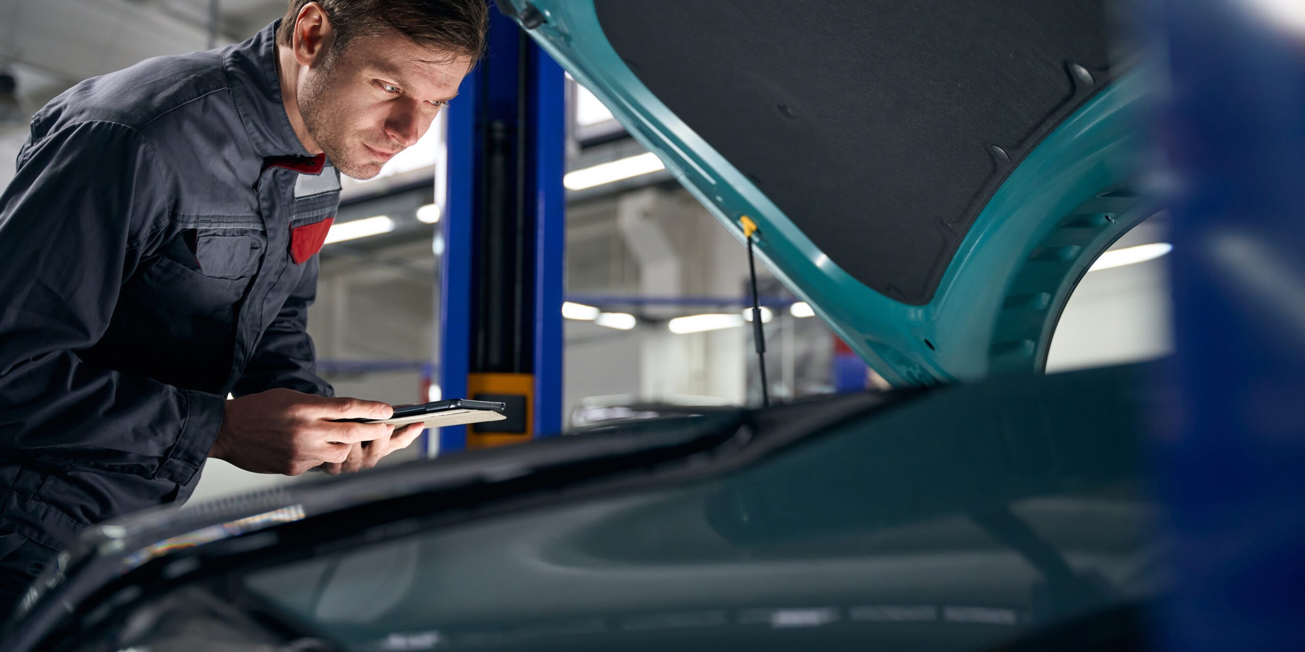 Male in protective clothes standing in tire fitting near car with open hood and holding laptop
